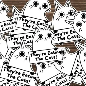 A bunch of stickers placed on a table of a white cat holding a sign that says, They're eating the cats