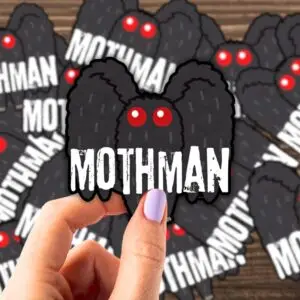 A hand holding a Mothman sticker over a table with a bunch of Mothman stickers on it